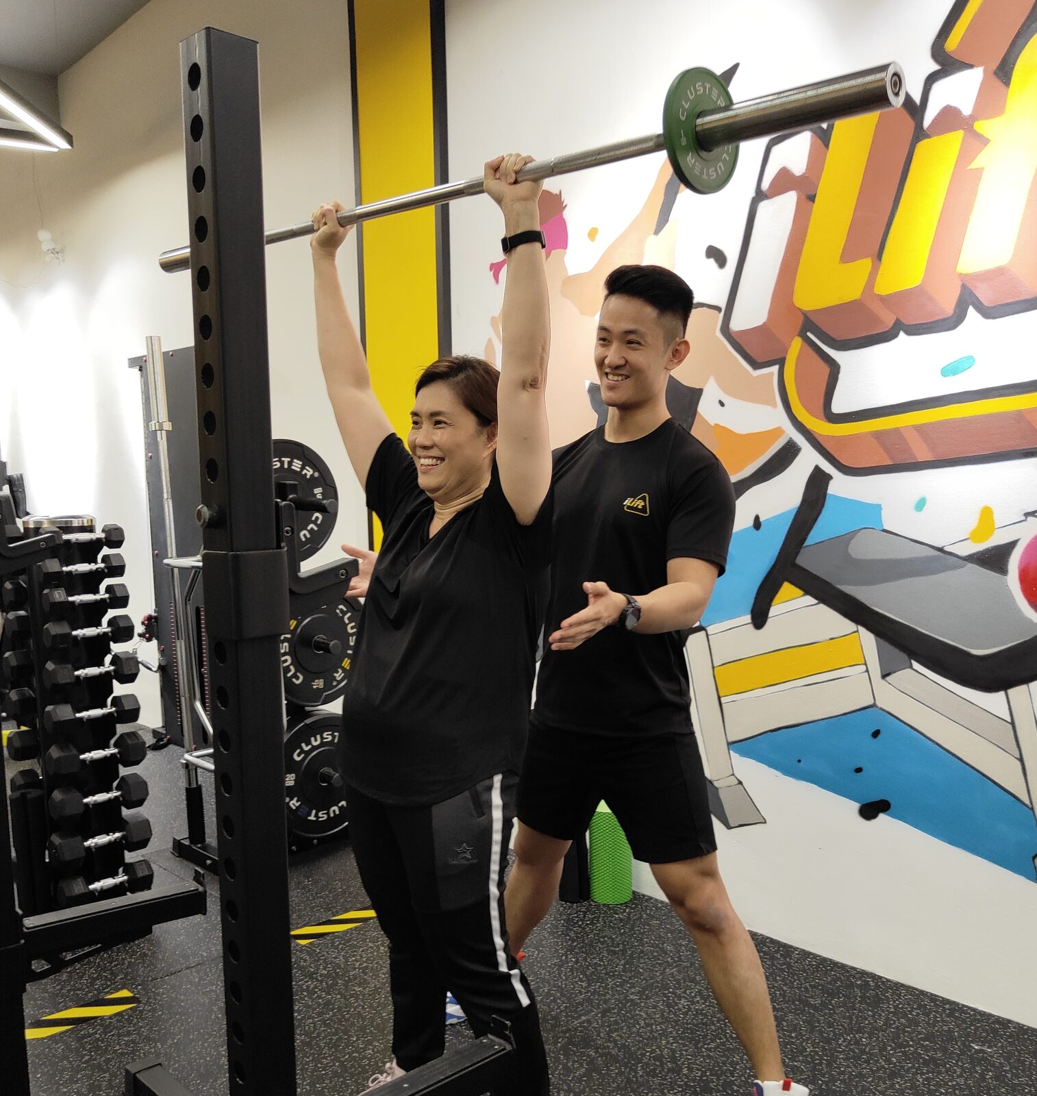 Coach guiding a client through an overhead barbell lift at iLift Gala City, Kuching