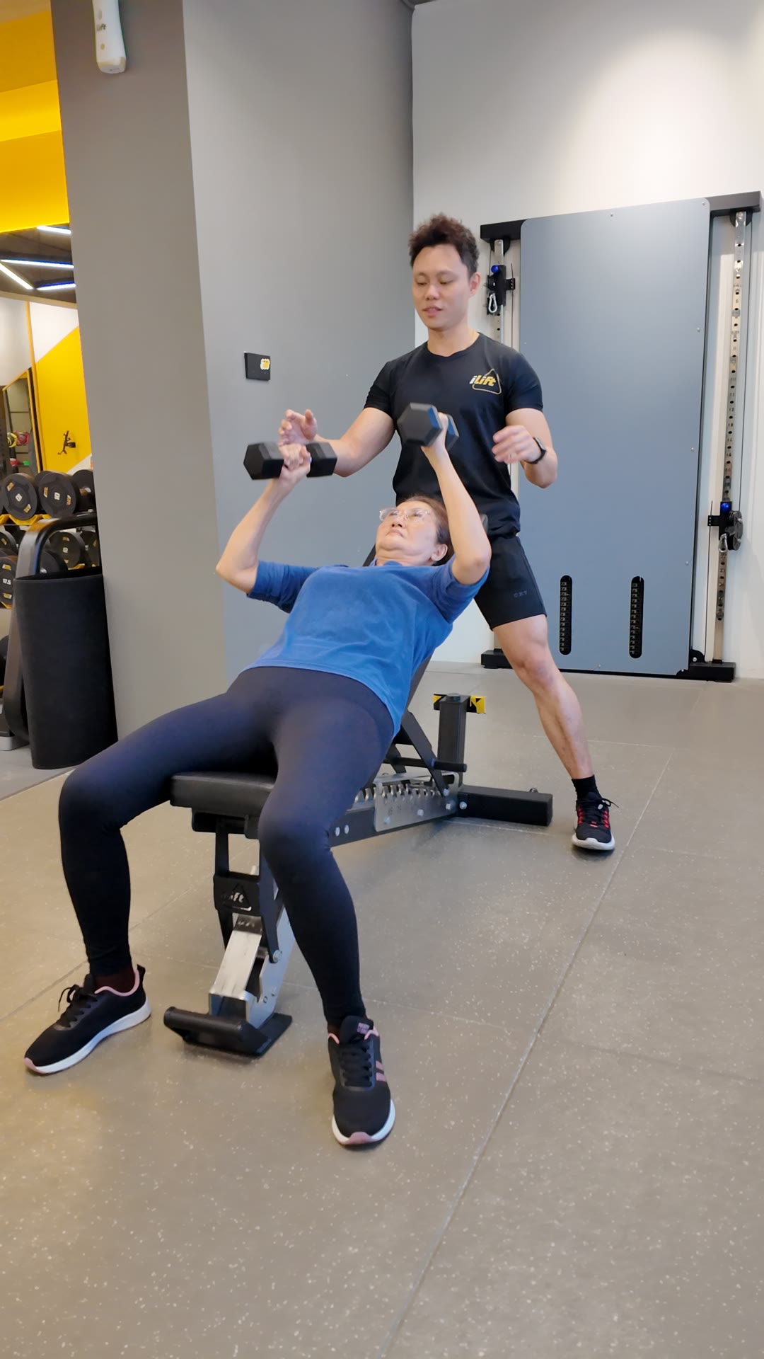 An older client doing an upper-body dumbbell press with her coach, a good option on a sore-knee day